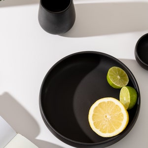 K&ouml;nnte beinhalten: Eine schwarze Keramiksch&uuml;ssel mit breitem Rand, gef&uuml;llt mit Zitronen- und Limettenscheiben. Die Sch&uuml;ssel steht auf einer wei&szlig;en Oberfl&auml;che mit einem Buch und einer schwarzen Keramikvase.