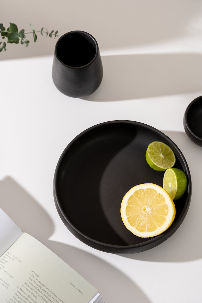 K&ouml;nnte beinhalten: Eine schwarze Keramiksch&uuml;ssel mit breitem Rand, gef&uuml;llt mit Zitronen- und Limettenscheiben. Die Sch&uuml;ssel steht auf einer wei&szlig;en Oberfl&auml;che mit einem Buch und einer schwarzen Keramikvase.