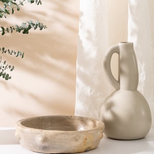 Könnte beinhalten: Eine hellbeige Keramikschüssel mit strukturiertem Rand und eine hellbeige Keramikvase mit rundem Körper und gebogenem Griff. Die Vase und die Schüssel stehen auf einer weißen Oberfläche mit einem weißen Stoffhintergrund.