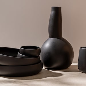 K&ouml;nnte beinhalten: Ein Satz schwarzer Keramikschalen und eine Vase, gestapelt und auf einer hellgrauen Oberfl&auml;che angeordnet. Die Vase hat eine einzigartige, bauchige Form.