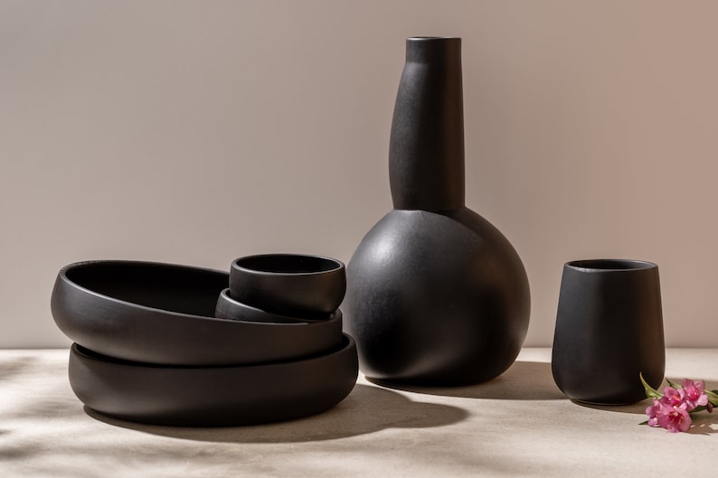 K&ouml;nnte beinhalten: Ein Satz schwarzer Keramikschalen und eine Vase, gestapelt und auf einer hellgrauen Oberfl&auml;che angeordnet. Die Vase hat eine einzigartige, bauchige Form.