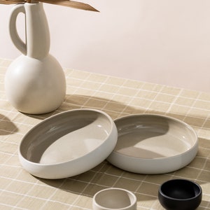 K&ouml;nnte beinhalten: Zwei gro&szlig;e, beige Keramik Schalen mit wei&szlig;en R&auml;ndern, eine kleinere beige Schale und eine schwarze Schale auf einer beige karierten Tischdecke.