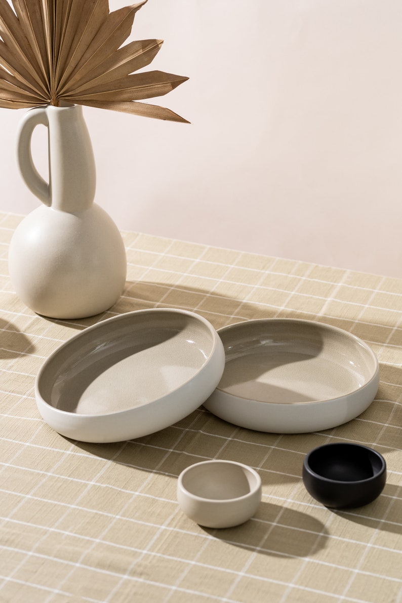 K&ouml;nnte beinhalten: Zwei gro&szlig;e, beige Keramik Schalen mit wei&szlig;en R&auml;ndern, eine kleinere beige Schale und eine schwarze Schale auf einer beige karierten Tischdecke.