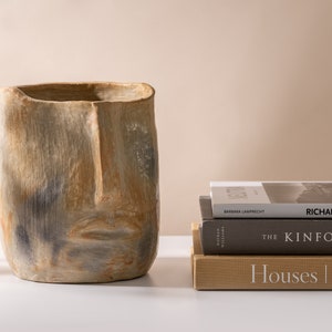 Peut inclure: Un pot en céramique en forme de visage avec un glaçage brun clair, gris et blanc. Le pot est posé sur une surface blanche devant une pile de trois livres. Le livre du haut porte le titre "Richard Neutra" sur le dos. Le livre du milieu porte le titre "The Kinfolk" sur le dos. Le livre du bas porte le titre "Houses | Atelier" sur le dos.