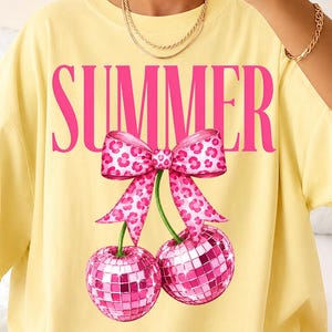 Op de afbeelding: Een lichtgeel t-shirt met het woord SUMMER in grote roze letters. Onder de tekst staat een afbeelding van twee roze discobal-kersen verbonden door een steeltje, bekroond met een roze strik met luipaardprint in het midden van het shirt.