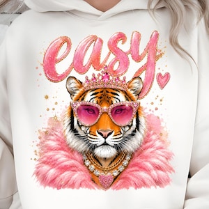 Könnte beinhalten: Ein cremeweißer Kapuzenpullover mit einem Grafikdruck eines Tigers, der eine rosa herzförmige Sonnenbrille, eine juwelenbesetzte Krone und eine rosa Federboa trägt. Das Wort easy ist in einer stilisierten rosa Schrift darüber gedruckt.