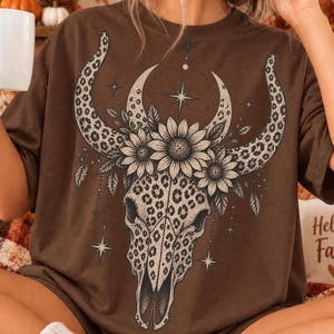 May include: A dark brown short-sleeve t-shirt featuring a large graphic print of a bull skull with a leopard print pattern. The skull is adorned with three sunflowers and surrounded by celestial star symbols and delicate leaf illustrations.