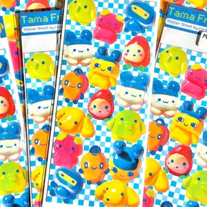 May include: A sticker sheet titled Tama Frens by ronilyn featuring a blue and white checkered background. The sheet contains multiple colorful, glossy, cartoon-style character stickers in various shapes, including round, square, and animal-like figures.
