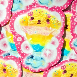 Può includere: Un adesivo fustellato raffigurante una pallina di gelato gialla a forma di animale all'interno di una coppa di vetro. Il dessert presenta strati blu e gialli, guarniti con accenti rosa e piccole stelle. Il design è bordato di rosa acceso.
