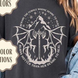Puede incluir: Camiseta gris oscuro con un diseño gráfico que presenta un hacha, alas e imágenes de montañas. El texto de la camiseta dice: "BEWARE THE FEMALE WHOSE DESIRE TO HURT BURNS HOTTER THAN HER DESIRE TO HEAL." La imagen también incluye el texto "COMFORT COLORS" y "6 COLOR OPTIONS."