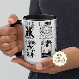 May include: White ceramic mug with a black handle and black rim. The mug features a black and gold design with the words "Fortos", "Lumos", "Umbros", and "Ignis" in a stylized font. Each word is accompanied by a graphic design.