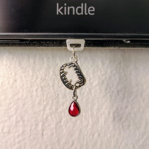 K&ouml;nnte beinhalten: Ein silberner Anh&auml;nger mit einem Fangzahn-Motiv und einem roten, tropfenf&ouml;rmigen Edelstein, der an einem wei&szlig;en Kunststoffclip befestigt ist. Im Hintergrund ist das Wort "kindle" zu sehen. Ein Gothic-Accessoire.
