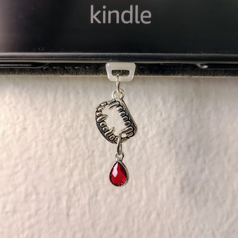 K&ouml;nnte beinhalten: Ein silberner Anh&auml;nger mit einem Fangzahn-Motiv und einem roten, tropfenf&ouml;rmigen Edelstein, der an einem wei&szlig;en Kunststoffclip befestigt ist. Im Hintergrund ist das Wort "kindle" zu sehen. Ein Gothic-Accessoire.