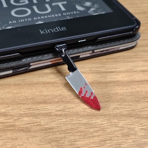 May include: A silver knife charm with a black handle and red blood splatter detail, hanging from a Kindle e-reader. The e-reader is in a patterned case, and the screen displays text from a book titled "Light Out".