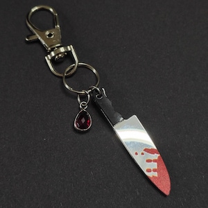 May include: A silver-colored knife-shaped keychain with a black handle and a blade stained with red. A small, teardrop-shaped red gem hangs from the keychain. The keychain has a silver clasp and a key ring.