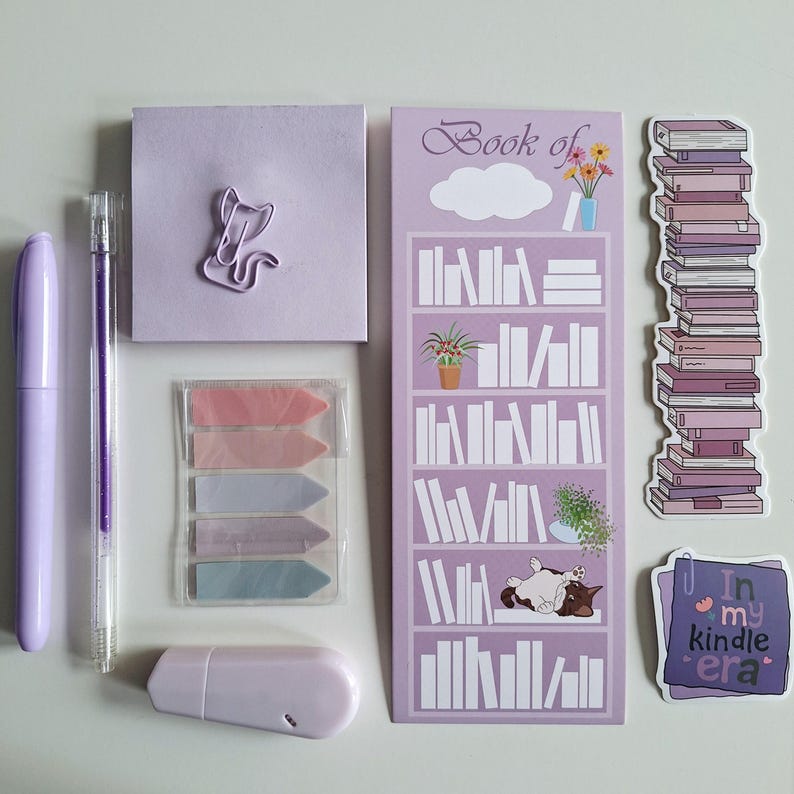 May include: A set of purple stationery supplies including a notepad, pens, sticky notes, a paper clip, a bookmark, and a sticker. The bookmark features a bookshelf with books and a cat. The sticker says "In my kindle era."