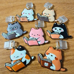 May include: A collection of seven enamel charms, each featuring a cartoon cat reading a book. The cats are depicted in various colors including light blue, beige, gray with glasses, pink, black and white, calico, and orange, all with clear plastic clips.