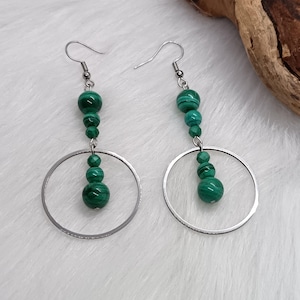Peut inclure: Une paire de boucles d'oreilles pendantes avec des crochets en métal argenté et de grands anneaux circulaires. Chaque boucle est ornée d'un alignement vertical de trois perles rondes en malachite verte aux rayures sombres, suspendues.