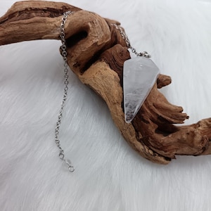 Può includere: Un pendolo di cristallo trasparente e sfaccettato con una catena d'argento. Il cristallo è sospeso a una catena e poggia su un pezzo di legno. Lo sfondo è una superficie morbida e bianca.