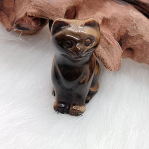 Op de afbeelding: Een kleine, gepolijste kattenfiguur, gemaakt van bruine en bruingele steen. De kat zit met opstaande oren. Het beeldje staat tegen een witte, donzige achtergrond en een stuk bruin hout.