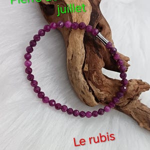Può includere: Un braccialetto circolare composto da piccole perle di rubino sfaccettate di colore rosa intenso con una chiusura cilindrica color argento. Il testo "Pierre de naissance de juillet" e "Le rubis" è stampato in verde e rosso sull'immagine.
