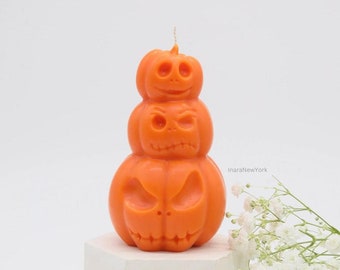 Op de afbeelding: Een gestapelde pompoen-kaars met drie oranje pompoenen, elk met een ander gesneden jack-o'-lantern gezicht. De kaars staat op een witte zeshoekige basis.
