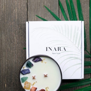 May include: A white coconut shell candle with a variety of colorful crystals and gold flakes embedded in the wax. The candle is in a wooden bowl and is surrounded by green leaves. The candle is labeled "INARA New York".