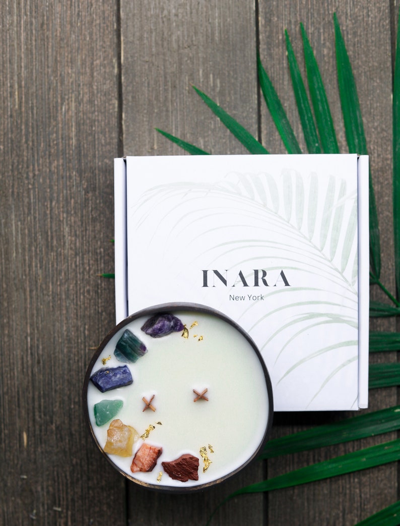 May include: A white coconut shell candle with a variety of colorful crystals and gold flakes embedded in the wax. The candle is in a wooden bowl and is surrounded by green leaves. The candle is labeled "INARA New York".