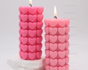 May include: Two pink heart-shaped pillar candles with flames burning. The candles are made of a soft, pink wax and are decorated with a pattern of hearts.