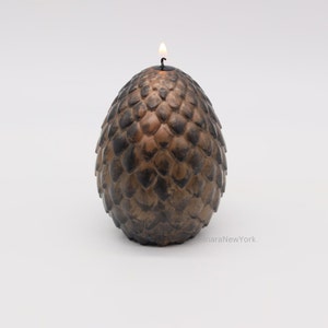 May include: A brown dragon egg-shaped candle with a lit wick on top. The candle is made to look like a dragon egg with textured scales.