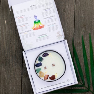May include: A white candle with seven different colored crystals embedded in the wax. The candle is in a white box with a description of the seven chakras and their corresponding crystals. The text reads "You deserve love, light and peace everyday of your life."
