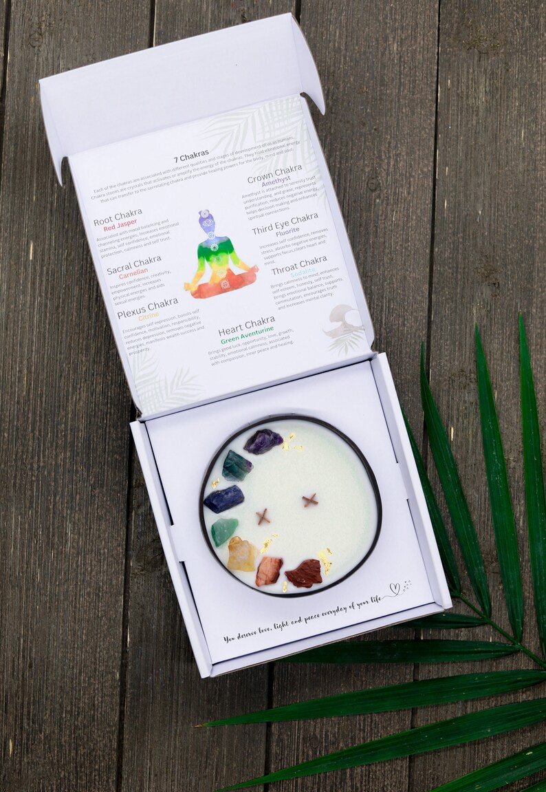 May include: A white candle with seven different colored crystals embedded in the wax. The candle is in a white box with a description of the seven chakras and their corresponding crystals. The text reads "You deserve love, light and peace everyday of your life."