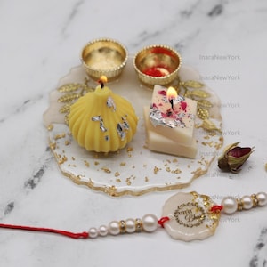 Puede incluir: Arreglo decorativo con una vela amarilla, una vela blanca y rosa, dos cuencos dorados y una pulsera de perlas con un cordón rojo. Los artículos están dispuestos sobre una bandeja blanca y dorada.