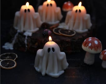 Könnte beinhalten: Weiße, geisterförmige Kerzen mit schwarzen Augen und brennenden Dochten. Die Kerzen sind auf einer dunklen Oberfläche mit dekorativen Pilzen und getrockneten Orangenscheiben angeordnet. Eine Halloween- oder herbstliche Dekoration.