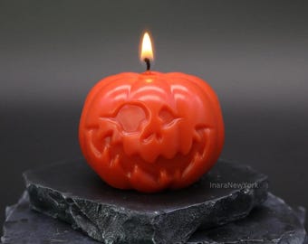Peut inclure: Une bougie rouge en forme de citrouille avec un visage de citrouille sculpté. La bougie est allumée et repose sur une pile de sous-verres en ardoise noire.
