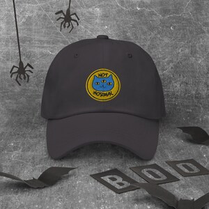 May include: Black baseball cap with a yellow embroidered patch that says "NOT NORMAL" and features a blue cat with three eyes.