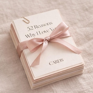 Könnte beinhalten: Ein Stapel Karten, gebunden mit einem hellrosa Satinband. Die oberste Karte trägt die Aufschrift "52 Gründe, warum ich dich liebe" mit dem Wort "KARTEN" darunter. Eine goldene Büroklammer sichert die obersten Karten. Der Hintergrund ist ein neutraler Stoff.