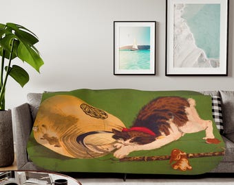 May include: A green throw blanket with a Japanese-style illustration of a cat with a red hat, lying on a large, round, tan object. The cat is looking down at the object.