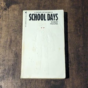 May include: A vintage paperback book titled "School Days: A Novel of Horror" by Robert Hughes. The cover features a white embossed illustration of a person and the title in bold black letters. The book is set against a dark wood grain background.