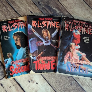 May include: Three vintage Fear Street books by R.L. Stine are displayed on a wooden surface. The book covers feature illustrations of young women and text, including "Cheerleaders" and "The Second Evil".