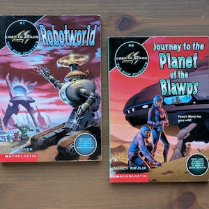 Puede incluir: Dos libros de bolsillo vintage de la serie "Lost in Space". El primero, "Robotworld", presenta una ilustración de robot. El segundo, "Journey to the Planet of the Blawps", muestra figuras con trajes espaciales. Ambos libros tienen portadas coloridas.