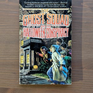 May include: A vintage paperback book titled "The Ghost Squad and the Halloween Conspiracy." The cover art shows children in costumes, a spooky house, and the text "Exciting mysteries peppered with humor." by E. W. Hildick.