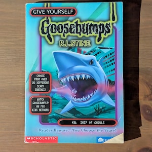 May include: A Give Yourself Goosebumps book titled Ship of Ghouls by R.L. Stine. The cover features a large blue shark with an open mouth, underwater scenery, and text including Choose from over 20 different scary endings and Reader Beware.