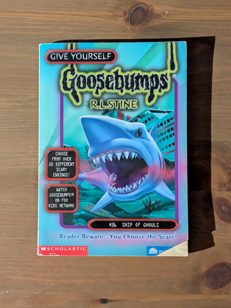 May include: A Give Yourself Goosebumps book titled Ship of Ghouls by R.L. Stine. The cover features a large blue shark with an open mouth, underwater scenery, and text including Choose from over 20 different scary endings and Reader Beware.