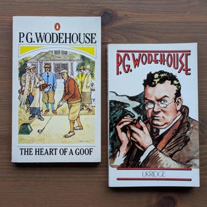 May include: Two vintage books by P.G. Wodehouse are displayed on a wooden surface. The book on the left is titled "The Heart of a Goof" and features a golf scene. The book on the right is titled "Ukridge" and features a portrait.