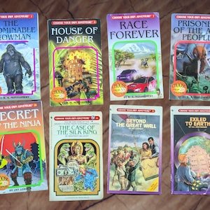 以下が含まれることがあります： 雪だるま、燃えている建物、レーシングカー、アリ、忍者、シルクの王、万里の長城、そして街を描いたカラフルな表紙の「自分の冒険を選ぼう」という本のセットです。本はすべて同じスタイルで、表紙に「自分の冒険を選ぼう」というタイトルが印刷されています。