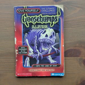 May include: A paperback book titled Give Yourself Goosebumps The Ultimate Challenge Special Edition 1 Into The Jaws Of Doom by R.L. Stine. The cover features a purple dinosaur skeleton illustration, red accents, and a Watch Goosebumps on Fox Kids Network badge.