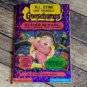 May include: A paperback book titled Give Yourself Goosebumps #20 Toy Terror: Batteries Included by R.L. Stine. The cover features a central illustration of a doll with pigtails in a pink dress, surrounded by toys, with purple holographic accents.