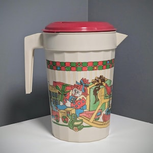 May include: A vintage cream-colored plastic pitcher with a red lid and handle. The pitcher is decorated with a colorful Christmas-themed design featuring a doll, presents, and a toy train. The pitcher has a capacity of approximately 64 ounces.