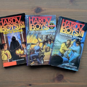 Puede incluir: Tres libros vintage de la serie Hardy Boys Casefiles se muestran sobre una superficie de madera. Las portadas de los libros presentan ilustraciones de escenas de suspense, incluyendo una figura sombría, un elefante y una lucha. Los títulos incluyen "Web of Horror", "Endangered Species" y "Rigged for Revenge".